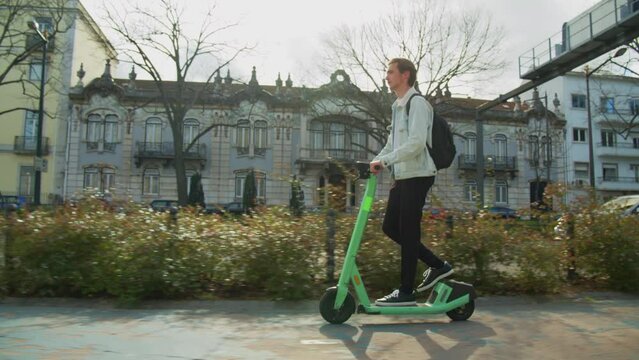 Young man rides an electric scooter in the city on a sunny day. Student riding on a rented green electric scooter through the streets of the city. Ecological transport. Slow motion