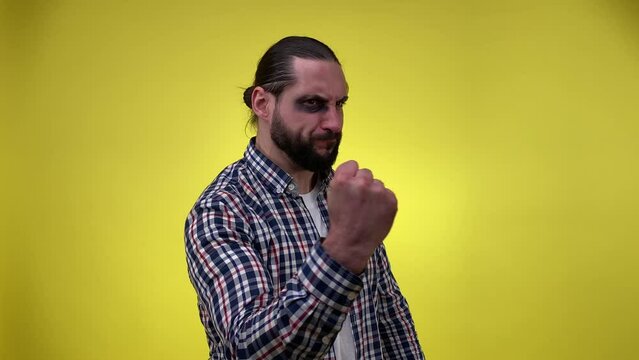 Bombastic mid adult man with black eye shaking his fist, threatening to sb. Bearded male person having eye injury after accident or fight with bruise. Indoors studio shot isolated on yellow background