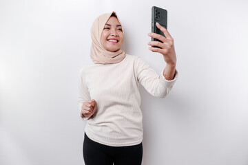 A happy Asian Muslim woman wearing a headscarf, holding her phone, isolated by white background