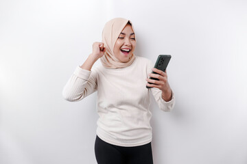 A young Asian Muslim woman with a happy successful expression wearing a hijab and holding smartphone isolated by white background
