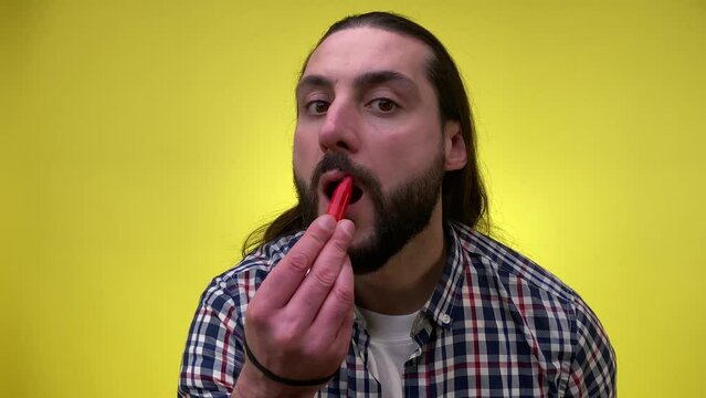 Mid adult male person with long hair wearing lip gloss. Bearded man puts make up on face, wearing lipstick, smacks lips looking at camera. Indoors studio shot isolated on yellow background. LGBTQIA