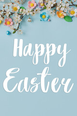 Happy Easter greeting card. Happy Easter text on candy flowers and blooming cherry branch flat lay on blue background. Modern Greeting card. Handwritten sign