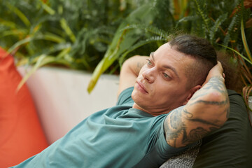 Pensive young man leaning back in armchair when resting in outdoor cafe