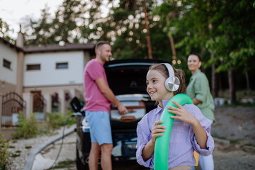 Happy family preparing for holiday, putting suitcases in car trunk, while their electric car charging.