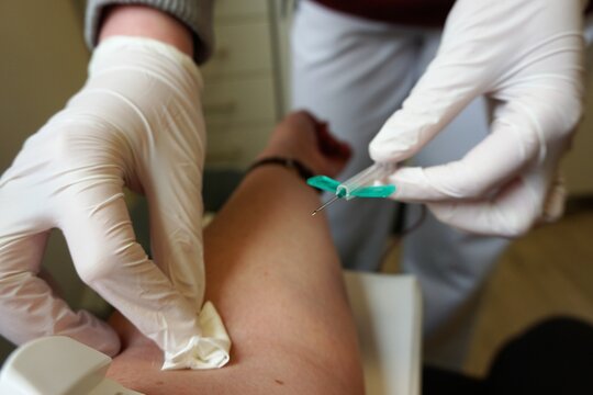 H&auml;nde von &Auml;rztin mit wei&szlig;en medizinischen Handschuhen h&auml;lt Infusionsnadel in linker Hand und h&auml;lt Wattetupfer auf Arm von Patientin nach Blutabnahme in Arztpraxis