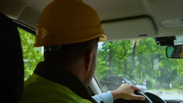 Back view of concentrated male engineer in helmet driving car and holding steering wheel on the way to the construction site. Real time video