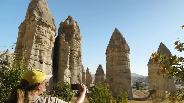 A blogger girl in a yellow baseball cap takes pictures of sandstone rocks in the form of phalos on a smartphone. Valley of Love in Cappadocia. Back view. Holidays in Turkey
