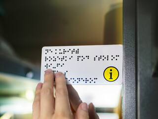 Hand of a person with blindness touches and reads Braille text plate with his hands on the door of...