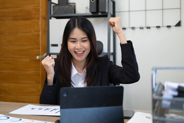Successful business woman with arms up celebrating. Excited asian woman sit at desk feel euphoric...