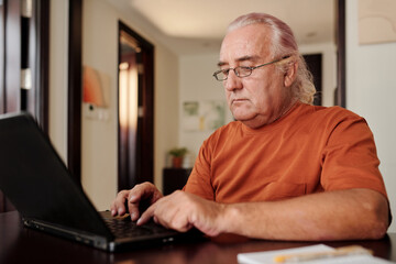 Senior man working on laptop at home, answering e-mails and filling documents