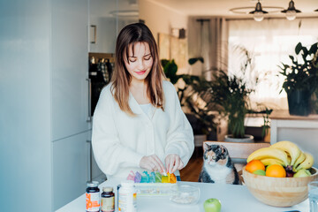 Woman sorting pills into pills organizer with her cat on the kitchen. Pill box with daily doses of medicines. Antioxidant diet vitamin supplements for health and beauty care, biohacking concept.