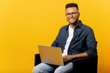 Nice smiling indian male entrepreneur using laptop sitting on the armchair isolated on yellow, portrait of happy determined male student looking at camera with computer on his lap, arab man typing