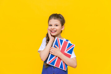 English for children. A little girl holds a textbook with an English flag in her hands. Children's language education. Yellow isolated background.
