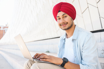Optimistic Indian handsome guy in red turban using laptop outdoors. Ethnic hindu freelancer man sitting on the steps in city landscape and looking at the camera, smiling friendly - obrazy, fototapety, plakaty