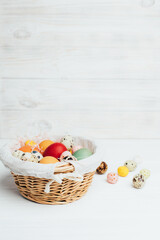 Colored eggs of different sizes in a straw basket on a wooden white background. Symbol of the Easter holiday. Easter background. Front view
