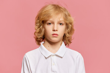 Portrait of little boy, child with curly blonde hair in white shirt attentively looking at camera against pink studio background. Concept of childhood, emotions, facial expression. Ad