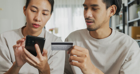 Happy young Asian couple sit on couch spend time together have fun use smartphone devices shopping use credit card online payment at home. Young husband and lifestyle domestic activity concept.