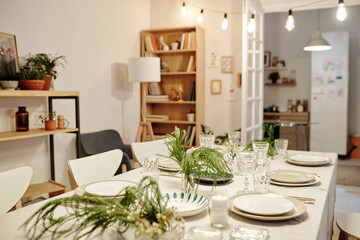 Part of living room with table served for family dinner with plates, wineglasses for drinks, green plants and candles used as decorations