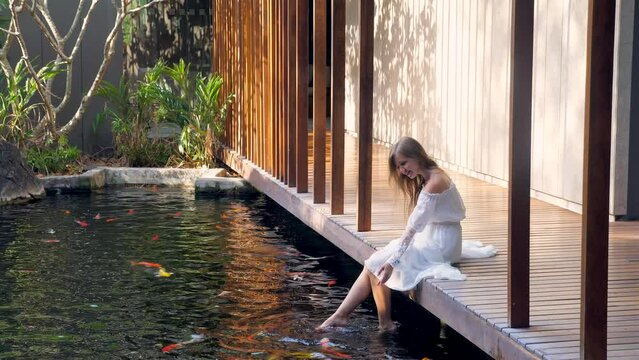 Woman relax in luxury spa with fish pond, enjoy bright morning and nature, relaxing and having good time sitting on wooden bridge barefoot. female playing feet in water at outdoor pond. Slow motion
