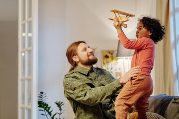 Happy young man in military uniform having fun with his cute little son playing with toy airplane while standing in front of him at home