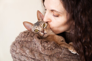 Close up of woman hugging and kissing her grey cat on a beige background. Cute Abyssinian kitten of blue color. Love relationship between human and cat. Cat day. Copy space. Selective focus.