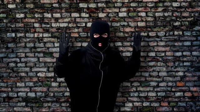 bandit puts hands up. Portrait of a thief wearing a black balaclava and gloves raising his hands up, isolated against a brick wall background.