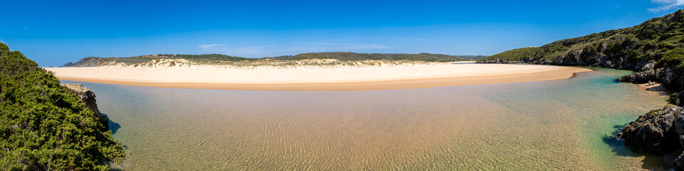 Crystal-clear river Ribeira de Aljezur with sand banks, dunes and a wide beach at Praia da Amoreira captured in an ultra-wide-angle panorama on a sunny day showcasing the beautiful nature of Portugal.