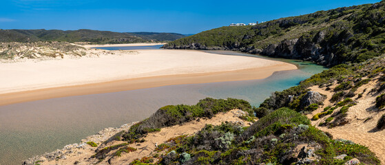 A stunning panoramic view of Praia da Amoreira shows a wide expanse of golden sand, crystal clear waters, and towering cliffs under a bright blue sky on a sunny afternoon in Portugal's Algarve region.