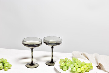 Gray shampagne glasses and grape om white wooden table, trend wine glass colored. Minimal summer still life with sparkling white wine in wide shampagne glass. Side view, copy space