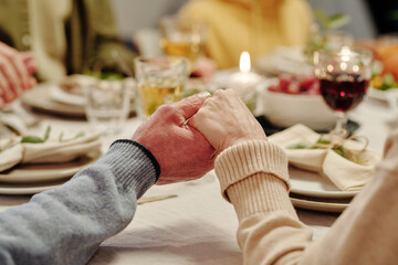 Hands of affectionate senior couple praying by served festive table with homemade food and burning...