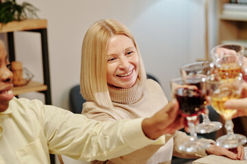 Happy blond mature woman clinking with wineglasses of guests sitting by festive table and enjoying family dinner in honour of life event