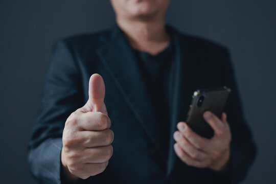 Businessman in suit showing thumbs up gesture and confidently holding mobile phone showing successful and confident, corporate leadership entrepreneur