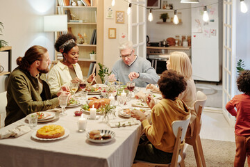 Members of large intercultural family sitting and communicating by served table while eating...