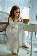 Little cute child girl in white nightie is embroidering with embroider frame at home.