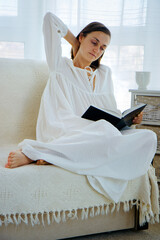 Portrait of the beautiful young woman in vintage nightwear is reading a book while sits on the sofa at home