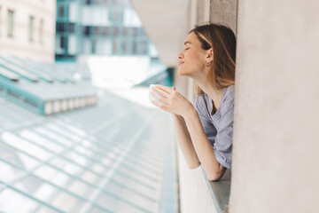 Fototapeta premium Young woman looking out the window and holding mug at home or hotel room. She drinking coffee or tea after wake up in the morning. Woman look enjoy and happy, positive mood, smell of morning coffee