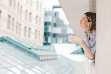 Young woman looking out the window and holding mug at home or hotel room. She drinking coffee or...