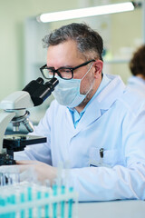 Mature expert in virology wearing lab coat, eyeglasses, protective mask and gloves studying cells of new virus in microscope in laboratory