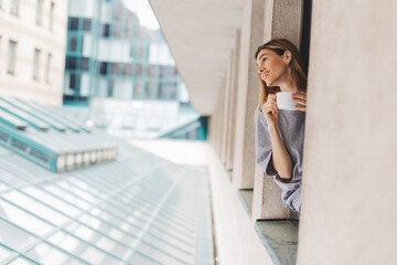 Young woman looking out the window and holding mug at home or hotel room. She drinking coffee or tea after wake up in the morning. Good morning! Woman look happy, positive mood, sitting on windowsill.