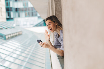 Young woman looking out the window and holding smart phone at home or hotel room. Woman using mobile phone chatting, video call or message, zoom in the morning. Woman look enjoy and happy, hi gesture.