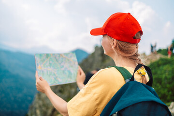 Back view of stylish woman holding paper map, wearing green backpack and red hat looking at mountain view while relaxing in nature. Travel and wanderlust concept. Amazing chill moment. High quality