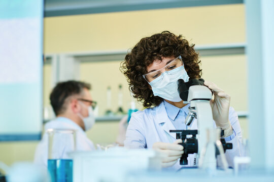 Mature Expert In Virology Wearing Protective Mask Adjusting Dial On Microscope While Studying New Virus In Clinical Laboratory