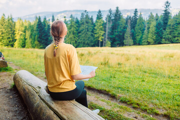 Back view of stylish woman holding paper map, wearing green backpack and red hat looking at mountain view while relaxing in nature. Travel and wanderlust concept. Amazing chill moment. High quality