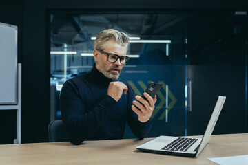 Senior serious gray-haired male businessman sitting in the office at the desk and using a mobile phone. Reads news, writes messages, checks mail.