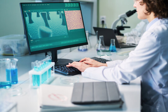 Young female researcher carrying out clinical experiment while sitting by desk in front of computer monitor with part of virus cell on screen