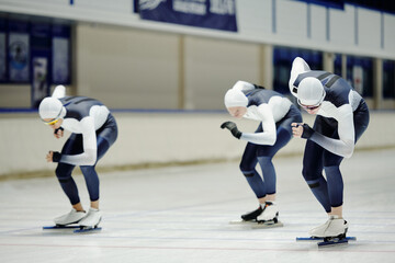 Fototapeta premium Group of fit youthful sportsmen in sports uniform sliding along ice rink in large modern arena taking part in short track speed skating race