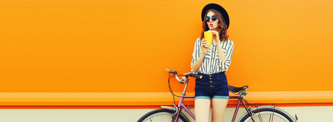 Summer colorful image of happy young woman drinking fresh juice with bicycle looking away on orange...