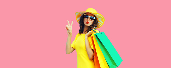 Portrait of beautiful happy young woman with shopping bags blowing her lips sending sweet air kiss wearing summer straw yellow hat on pink background