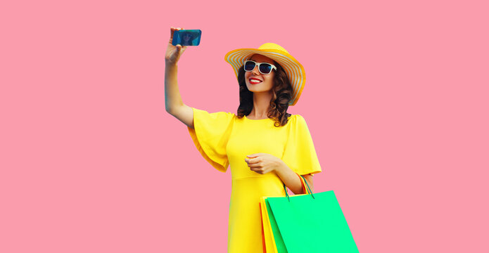 Beautiful happy smiling woman taking selfie by smartphone with shopping bags wearing yellow dress, summer straw hat on colorful pink background