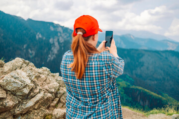 Young woman relaxing on the top mountains, holds a mobile phone in her hands and takes pictures of the landscape. Back view.